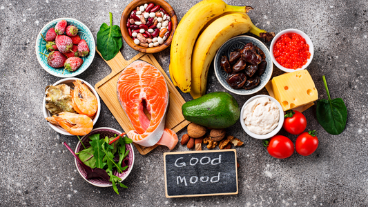 Healthy foods for yeast infection diet including salmon, bananas, avocado, nuts, tomatoes, shrimp, leafy greens, cheese, and berries arranged on a gray surface with a "Good Mood" chalkboard sign.