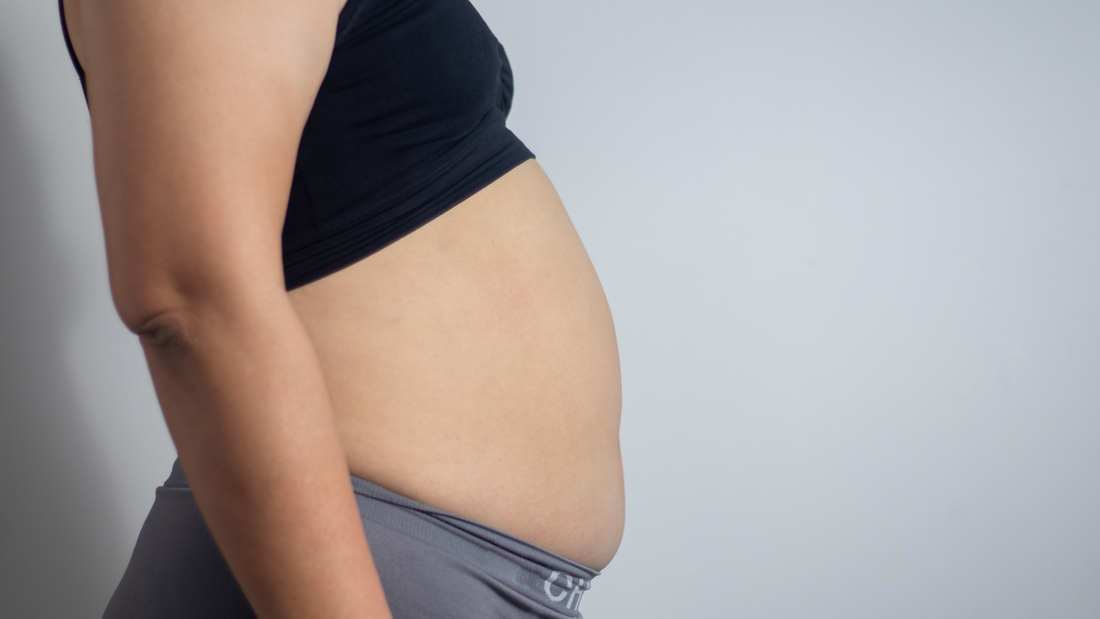 side view of woman with stomach bloating wearing black top and gray pants against plain background
